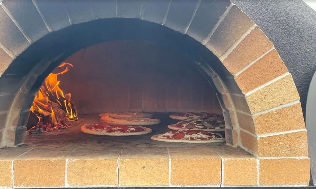 Pizzas cooking on the deck of a wood-fired oven beside an open oak fire